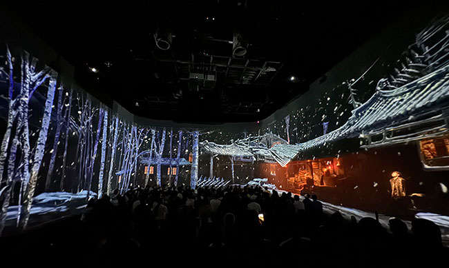 A tree and snow-filled landscape is projected onto three walls of a room while a group of people look on. 